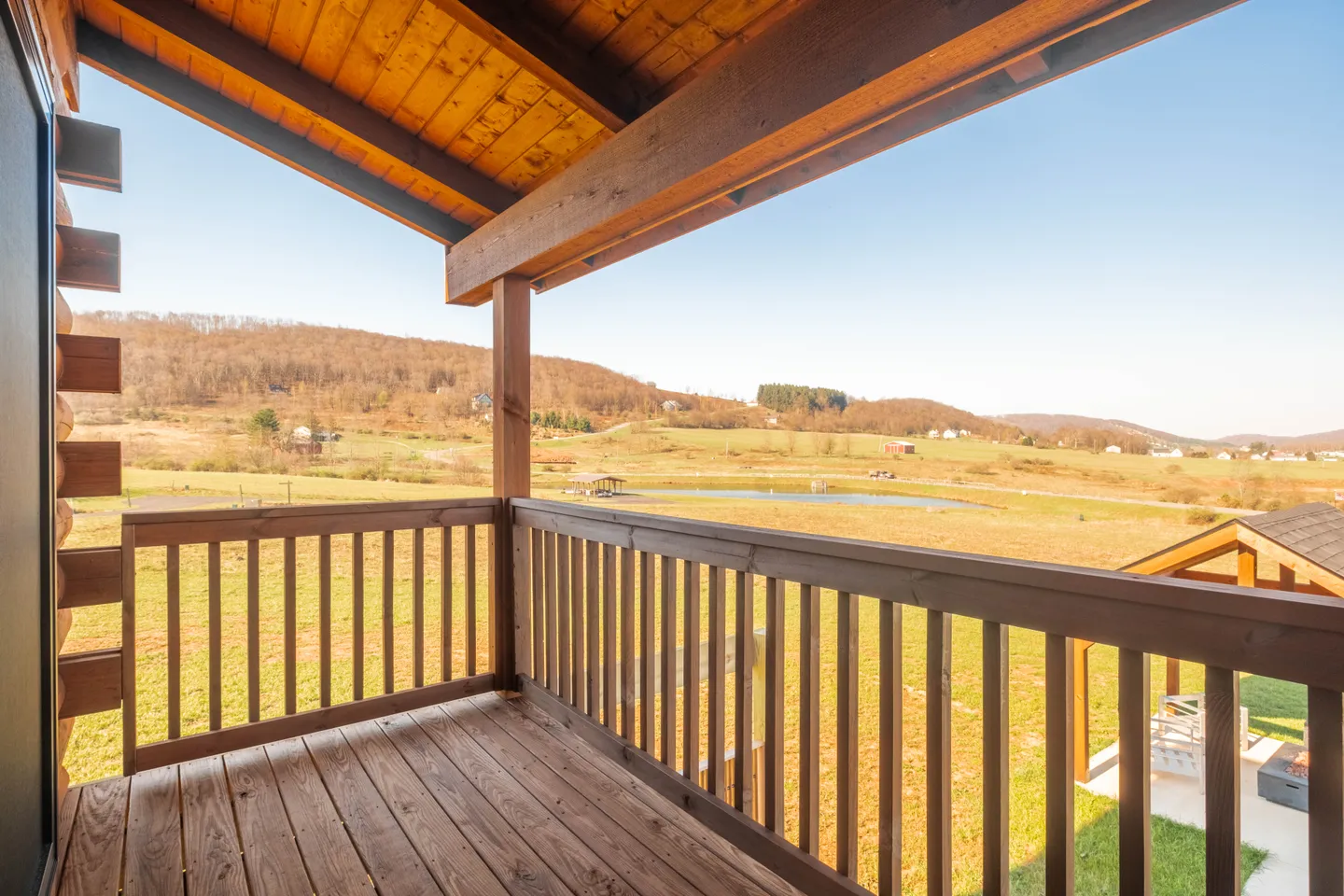 A covered wooden balcony overlooking an open field, a pond, and rolling hills under a clear blue sky.