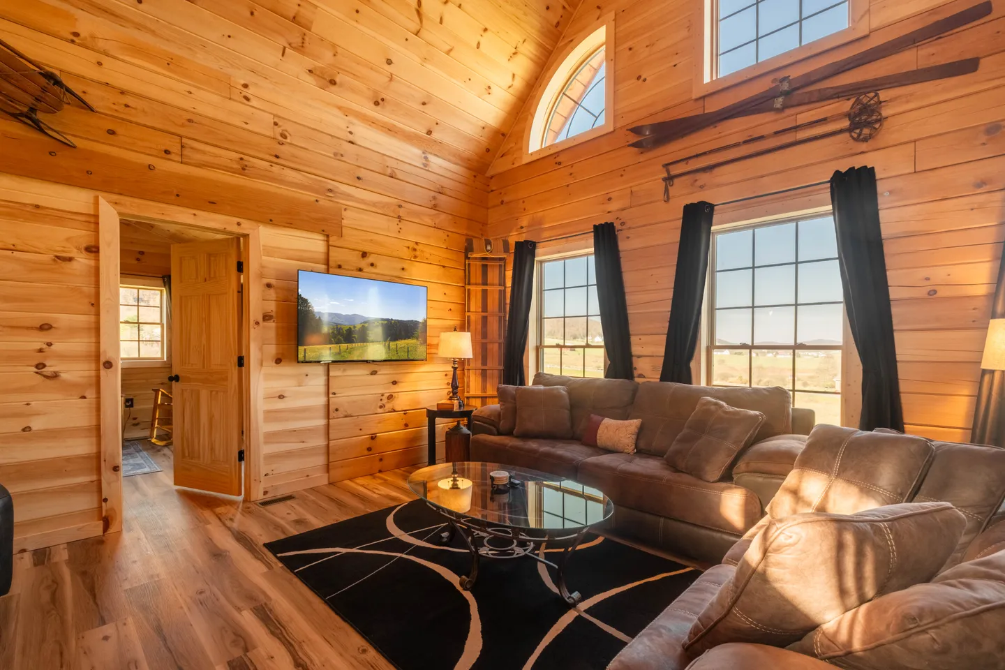 Living room with knotty wood-paneled walls and ceiling, a brown sectional sofa, glass coffee table, wall-mounted TV, and large windows.