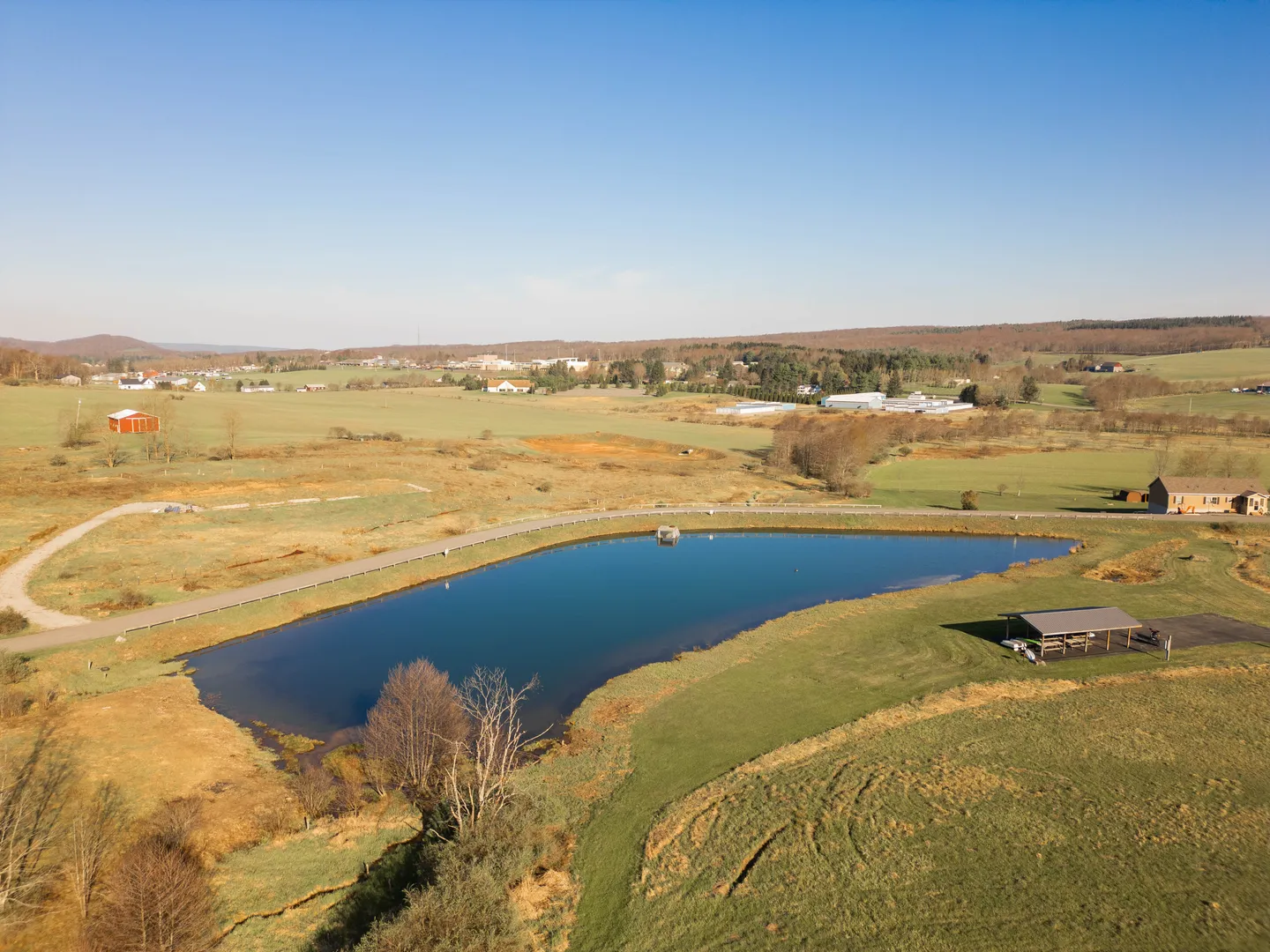 Aerial view of a rural property featuring a large pond, a small dock, a covered pavilion, and surrounding grassy fields.
