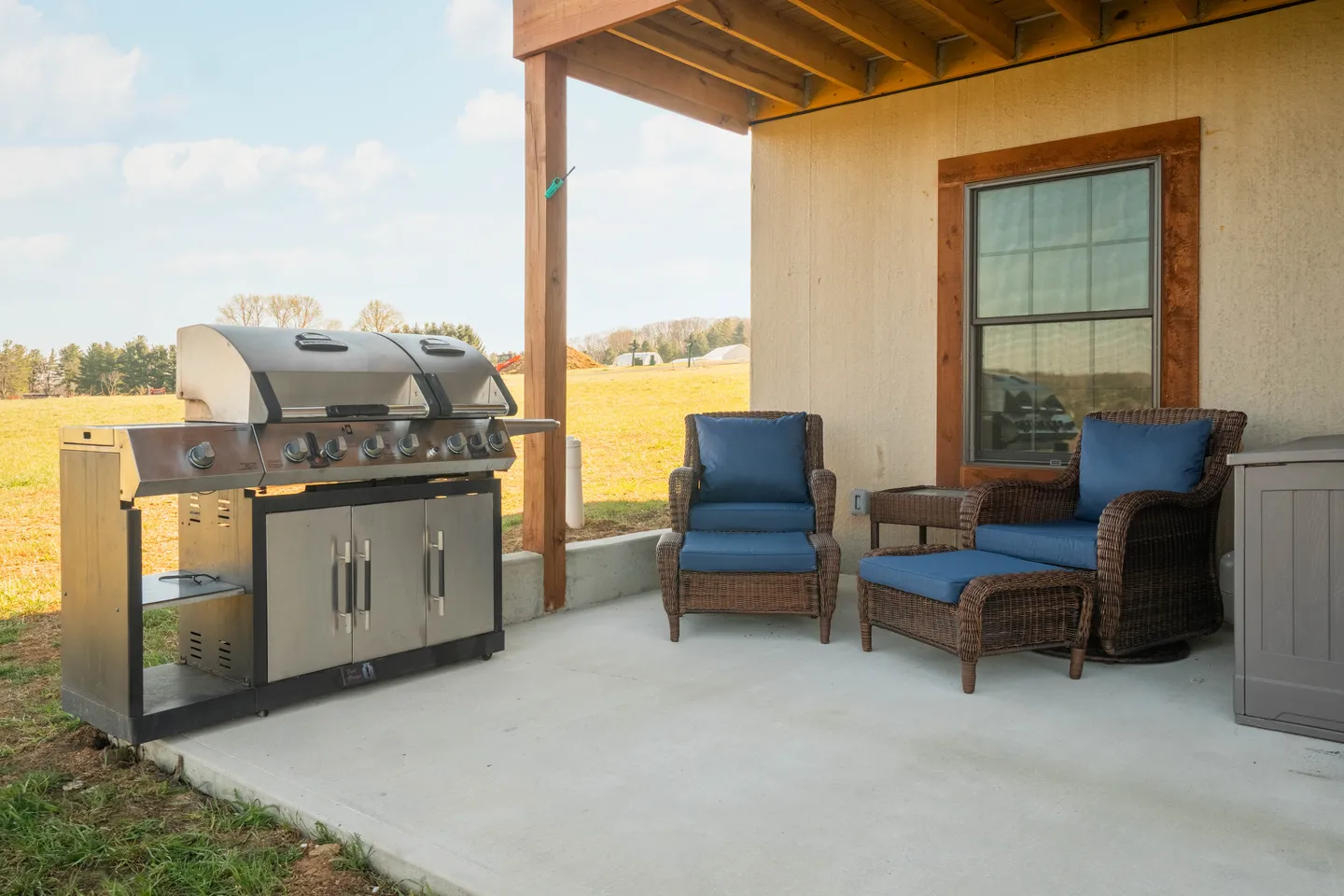Covered patio featuring a large stainless steel barbecue grill, wicker armchairs with blue cushions, and an ottoman facing an open field.