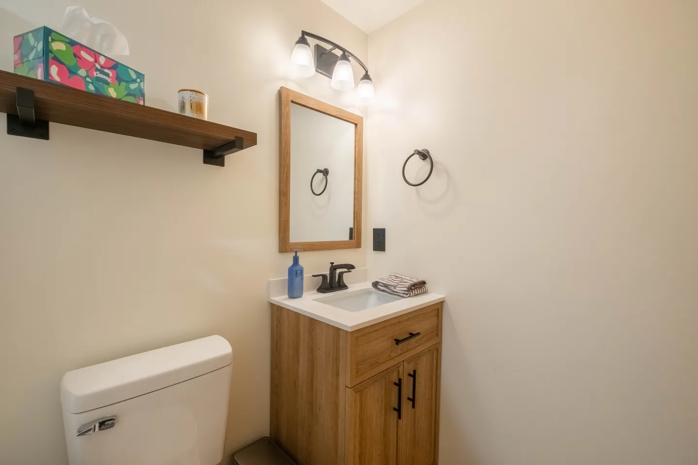 Bathroom with a light-toned wooden vanity, framed mirror, light fixture, toilet, and an open floating shelf.