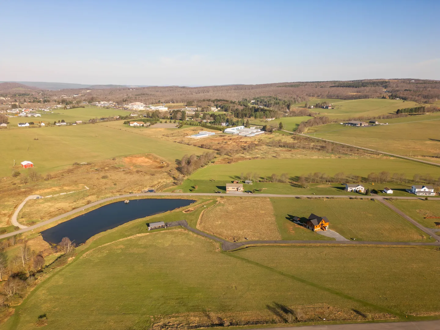 High aerial view of a rural landscape with open green fields, a private pond, and a single house with an outbuilding beneath a clear sky.