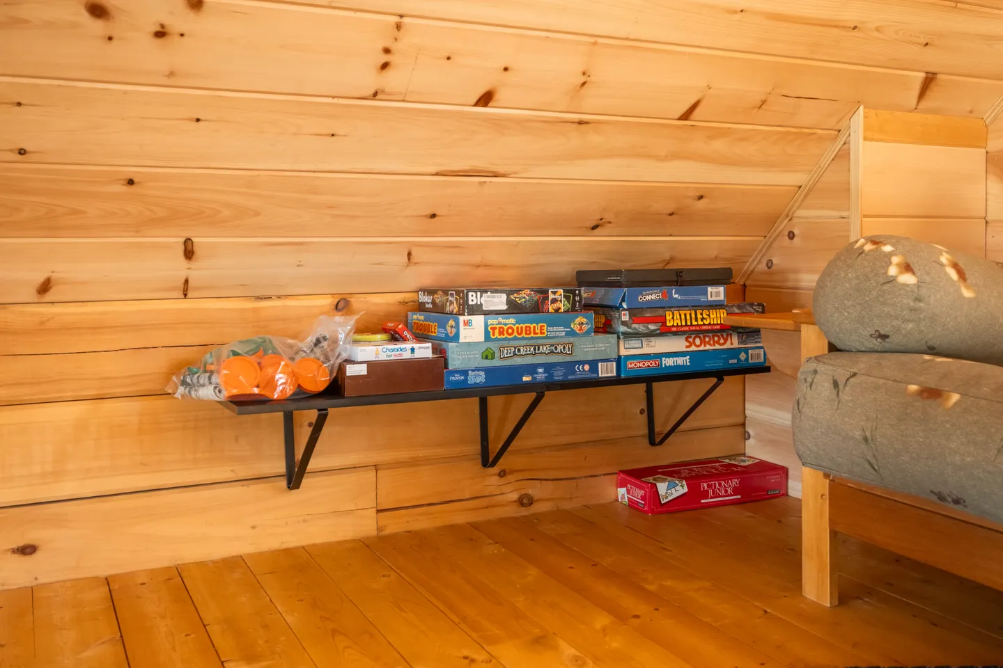 A wall-mounted black shelf lined with board games beneath a sloped pine wood ceiling next to a bed.