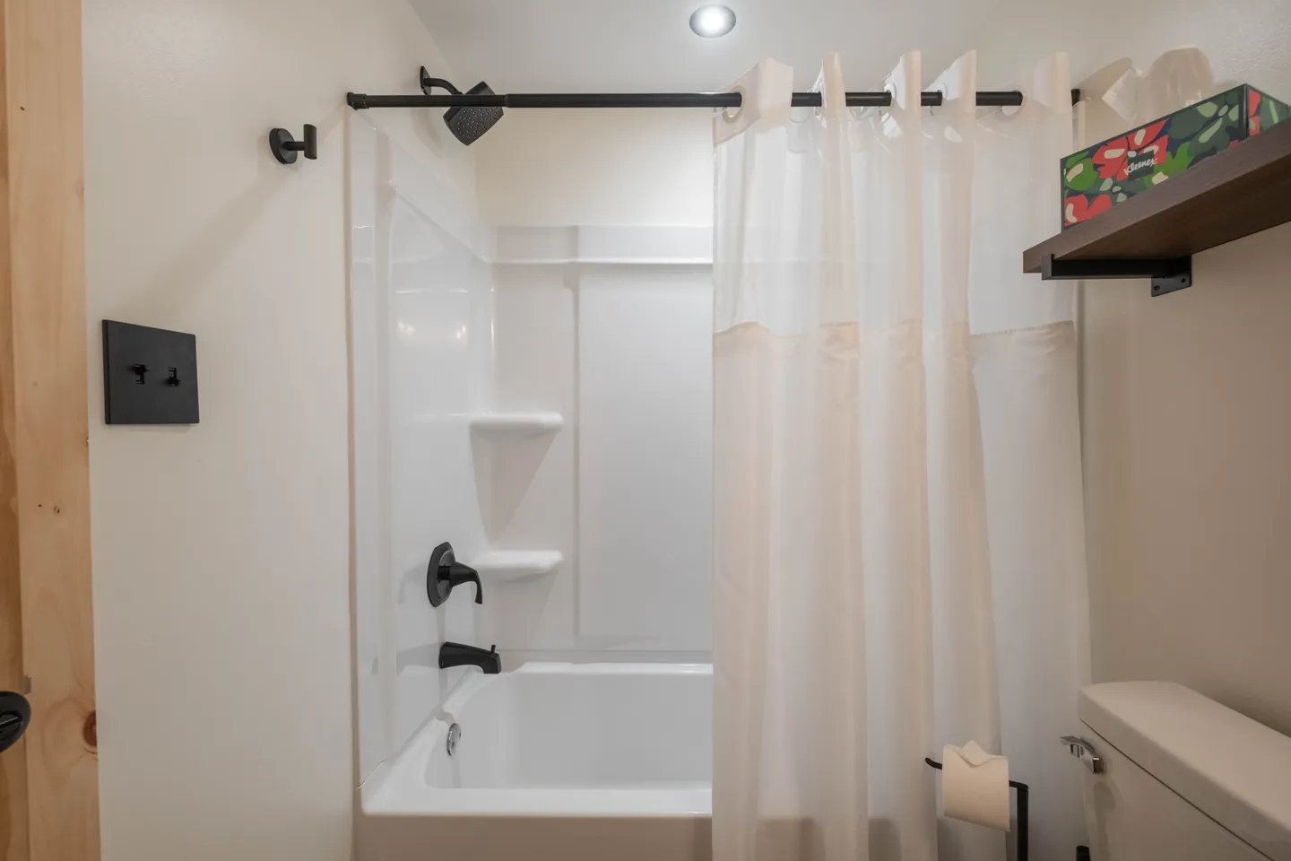 Bathroom showing a white bathtub and shower surround with dark fixtures, white shower curtain, and a partial view of the toilet.