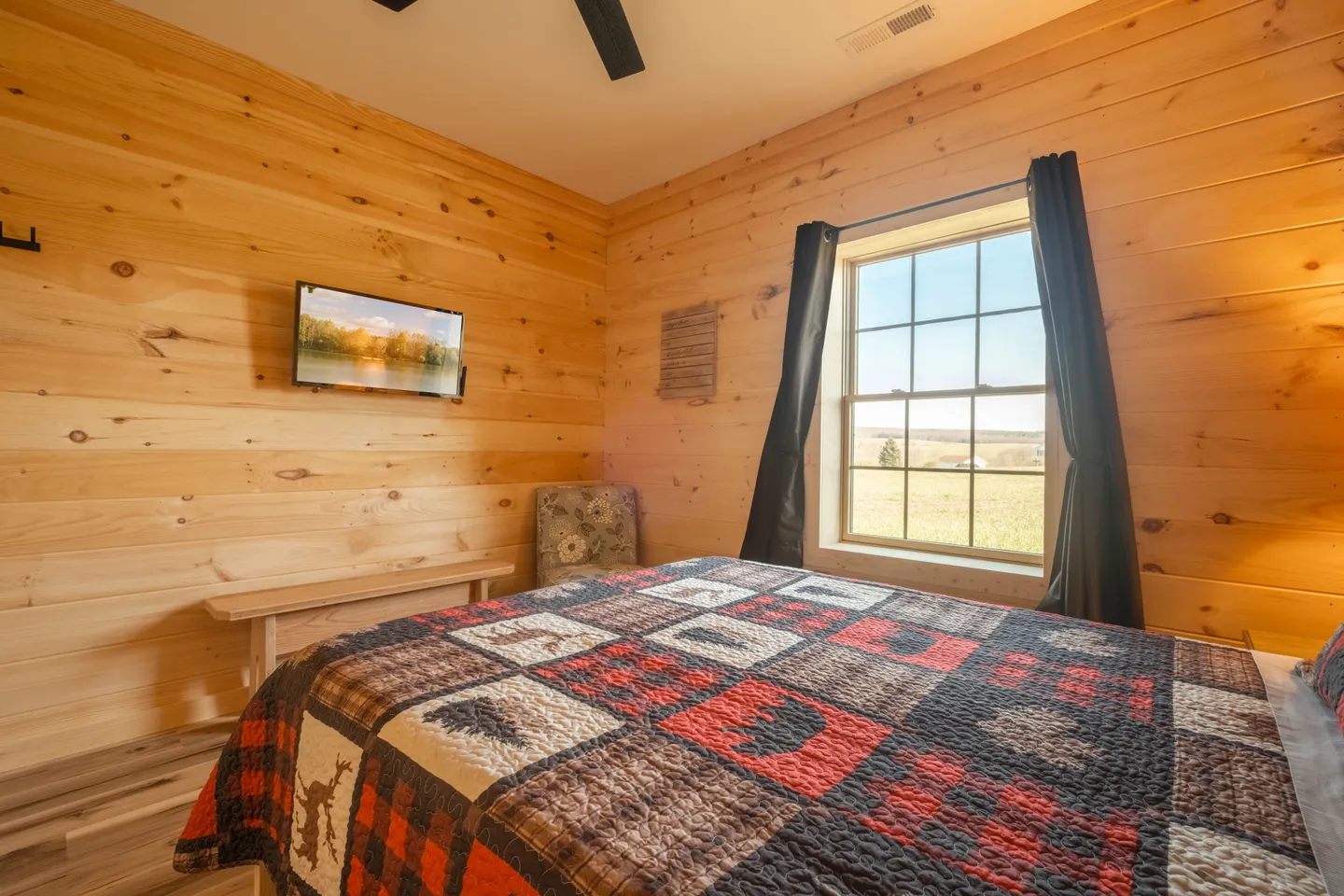 Bedroom with light wood paneled walls, a bed with a rustic patchwork quilt, a wall-mounted TV, and a window with views of an open field.