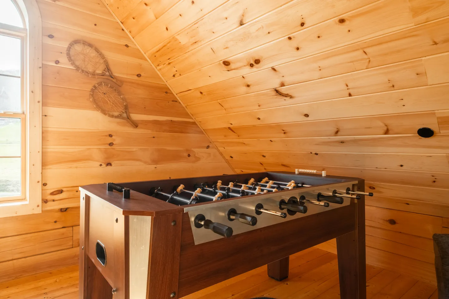 A foosball table situated under a sloped wooden ceiling with wood-paneled walls and snowshoes displayed as decor.