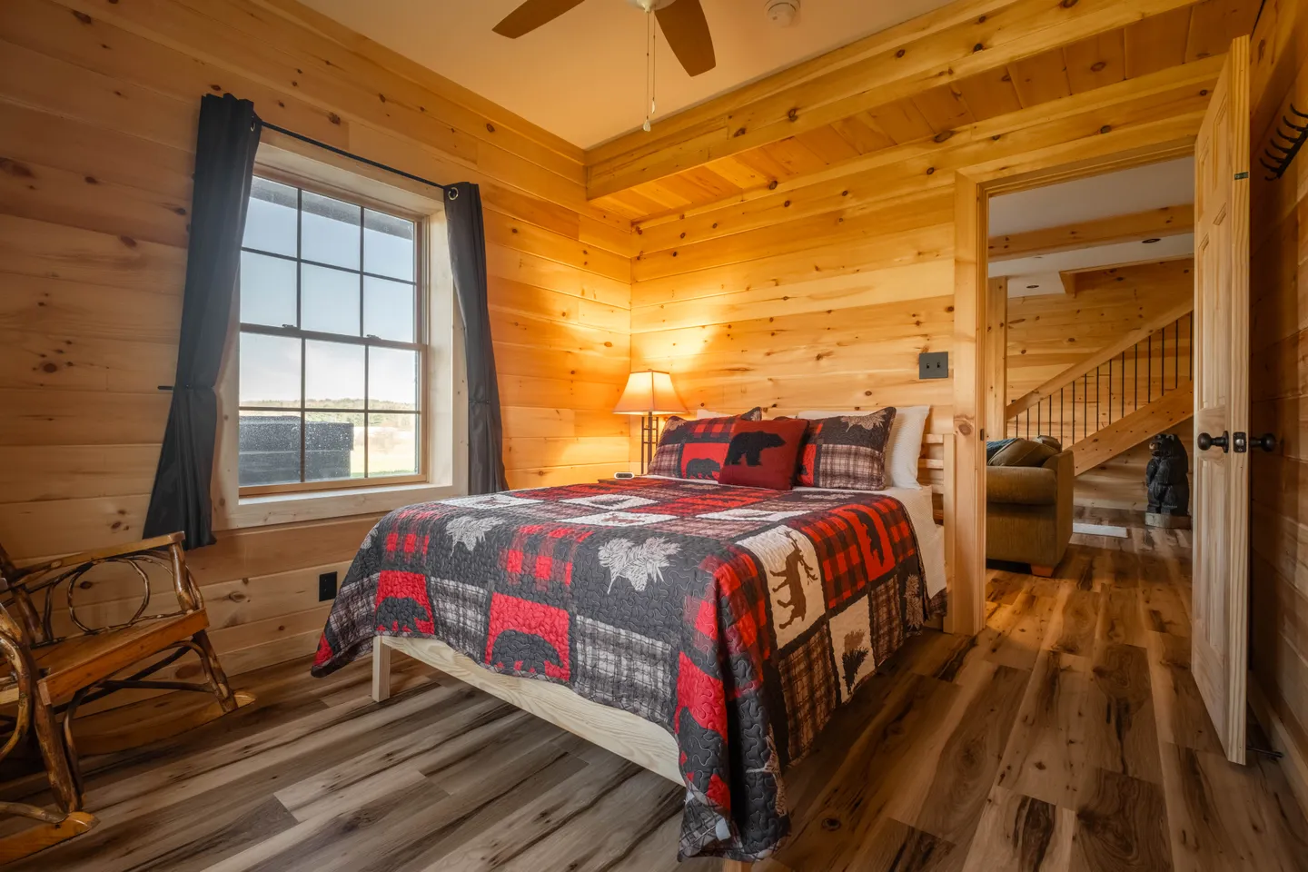 Bedroom with wooden plank walls and floors, featuring a bed made with a rustic patterned quilt and a rocking chair near the window.