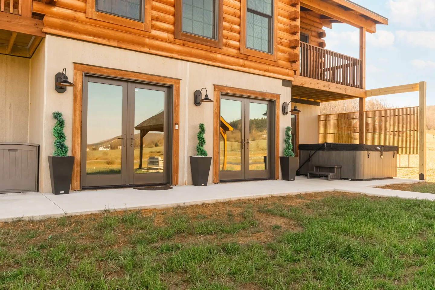 Lower patio level of a log cabin with two double glass doors, potted plants, and a covered hot tub.