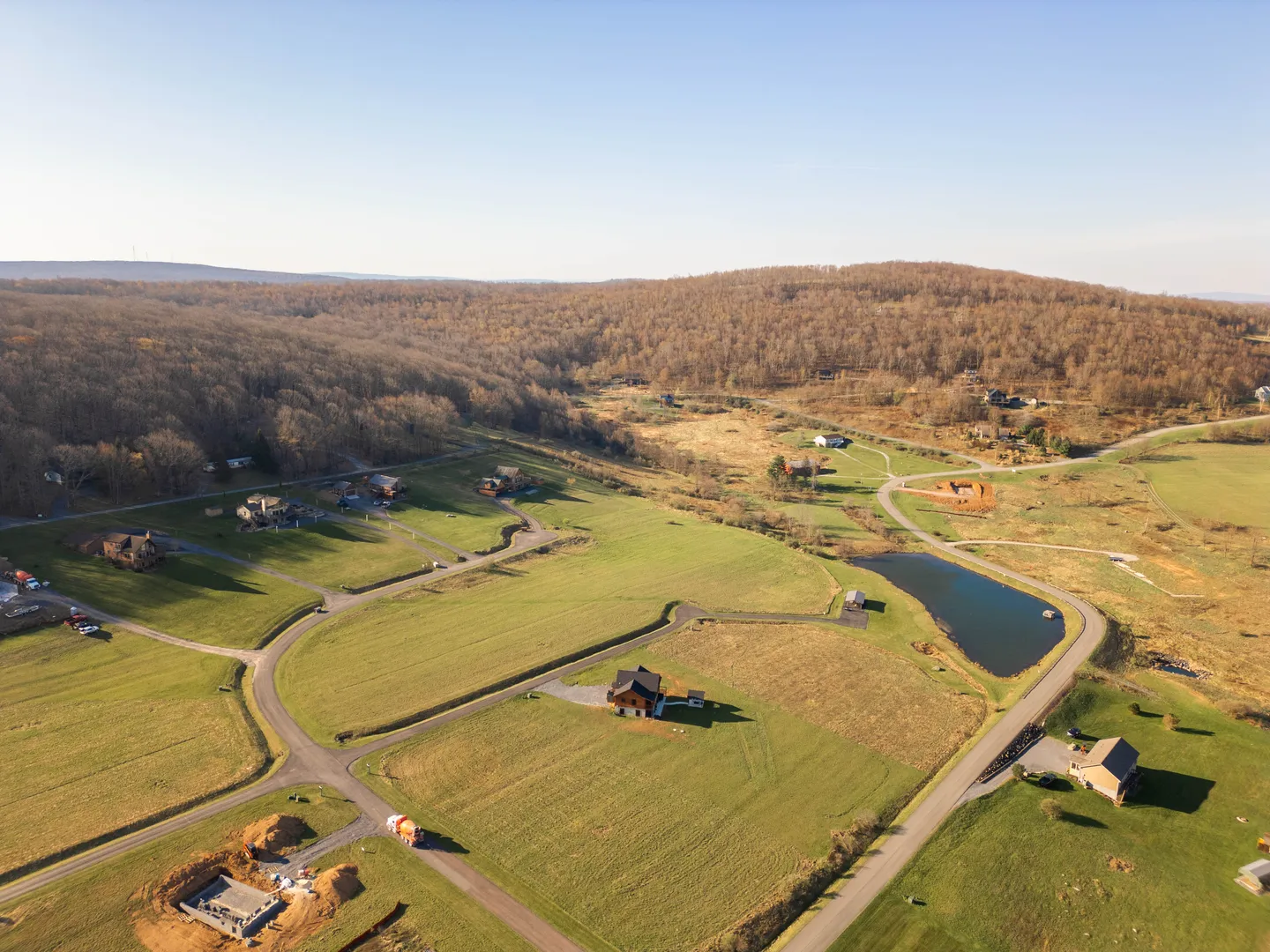 Aerial view of a rural residential development with scattered houses, paved roads, open grassy fields, a small pond, and wooded hills under a clear blue sky.