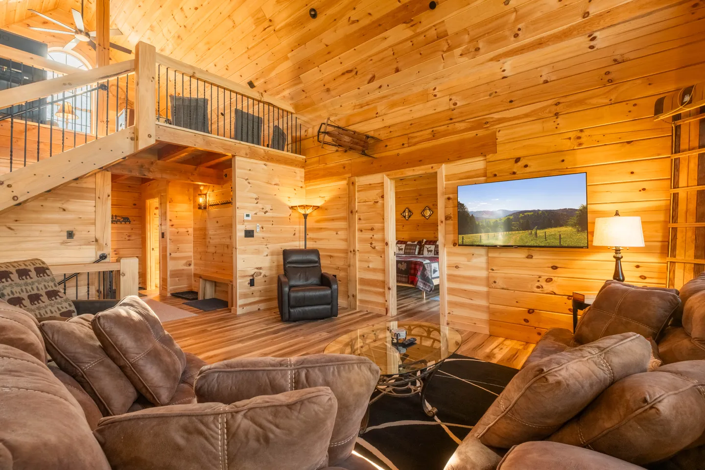 Living room with knotty pine wood walls, reclining sofas, a glass-top coffee table, wall-mounted television, and a staircase leading to a loft.