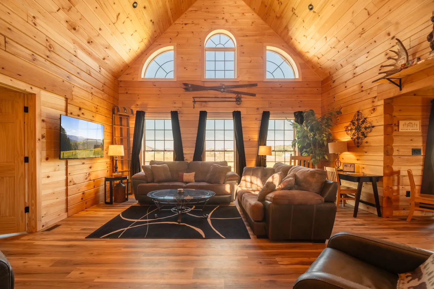 Living room with wood-paneled walls and vaulted ceiling, plush sofas, area rug, and tall windows.