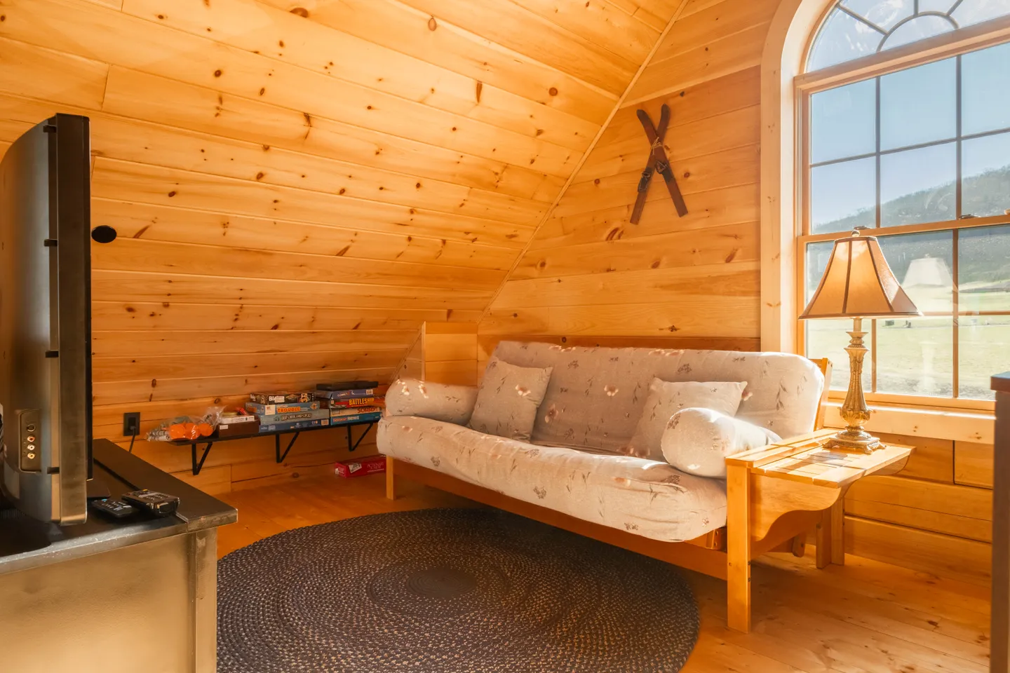 Wood-paneled loft area with a futon couch, round rug, television, and an arched window looking outside.