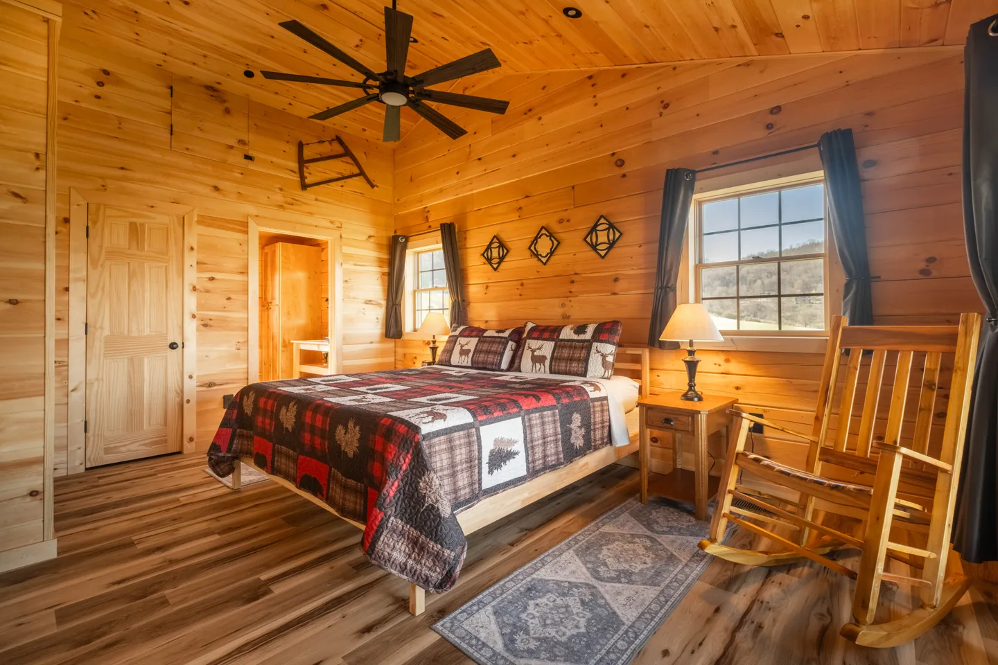Rustic wooden bedroom with wood-paneled walls and ceiling, a bed with a patchwork quilt, a rocking chair, and two windows.