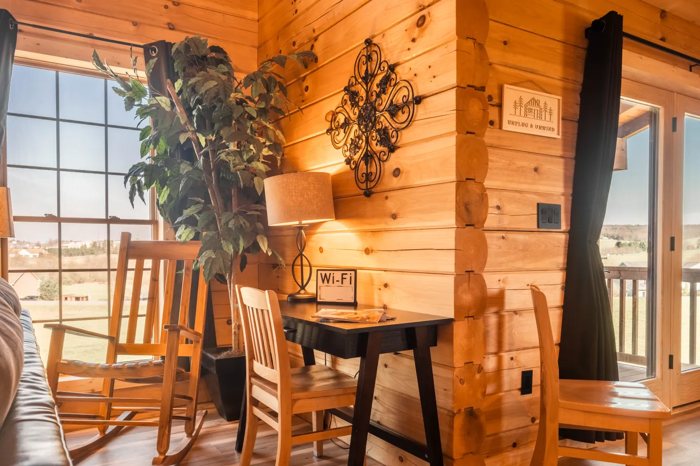 A corner of a log cabin room featuring a wooden rocking chair, a small desk with a lamp, a faux potted tree, and large windows looking outside.