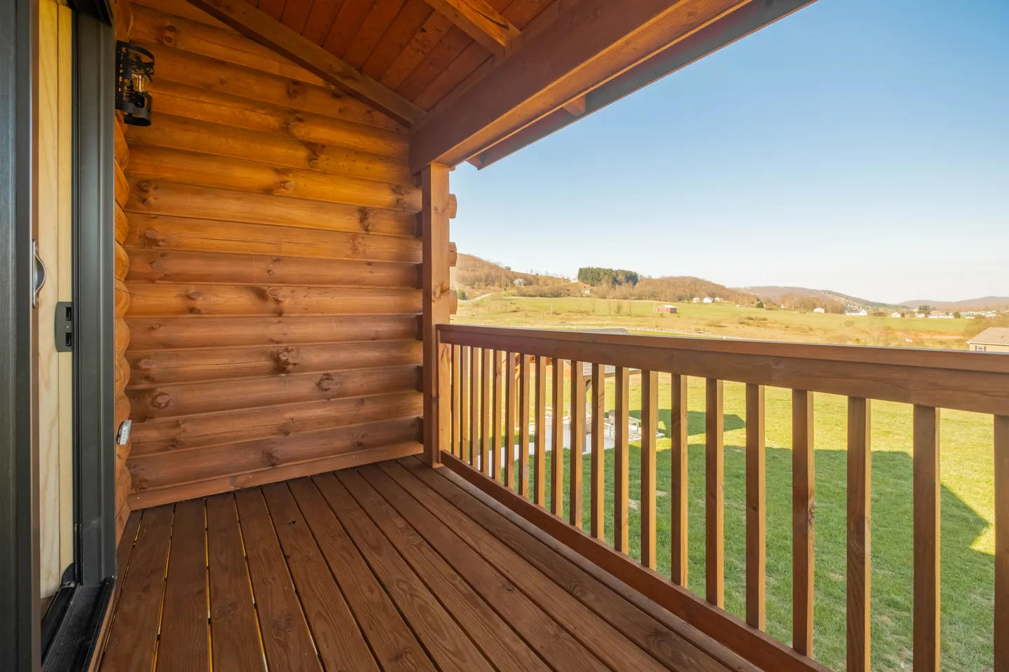 A covered wooden balcony on a log cabin with views of rolling green hills under a clear blue sky.