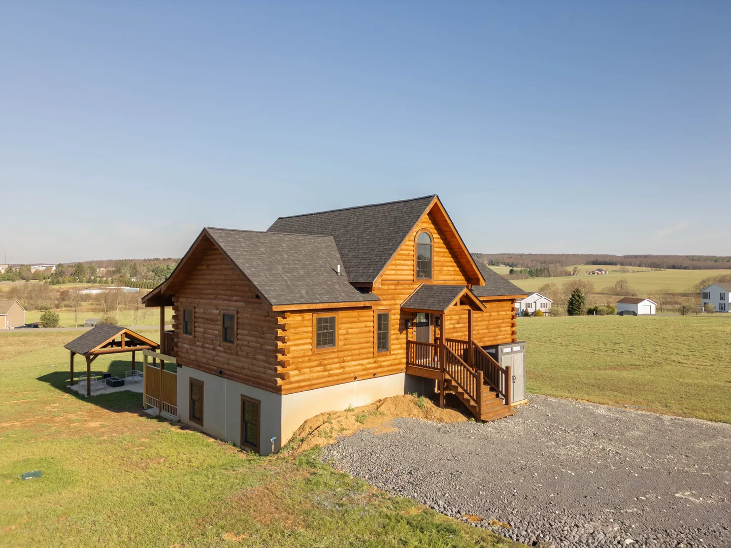 Exterior view of a multi-level log cabin home on a grassy lot with a gravel driveway and small detached pavilion.