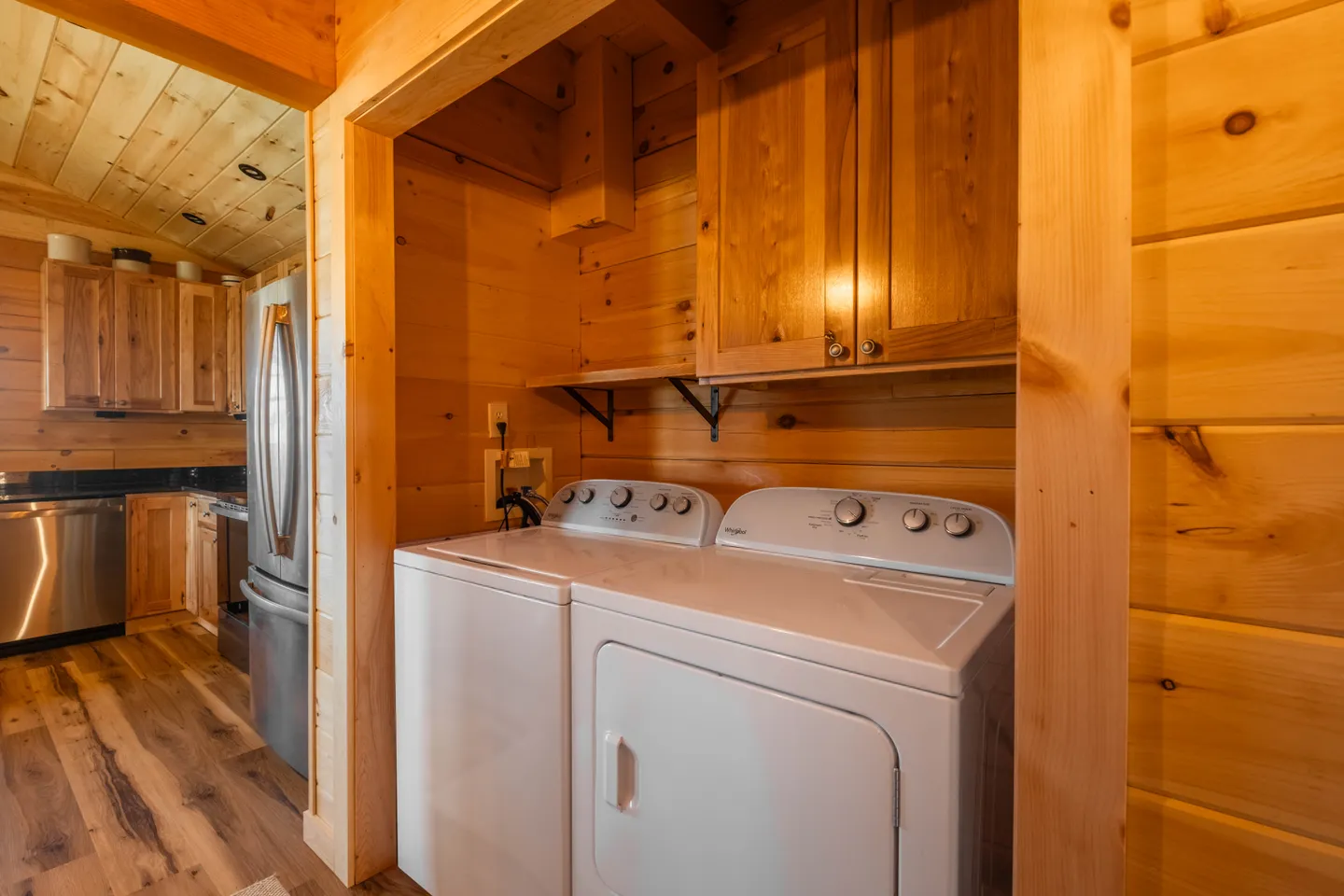 A laundry alcove with a white washer and dryer under wood cabinets, adjacent to a kitchen with wood paneling.