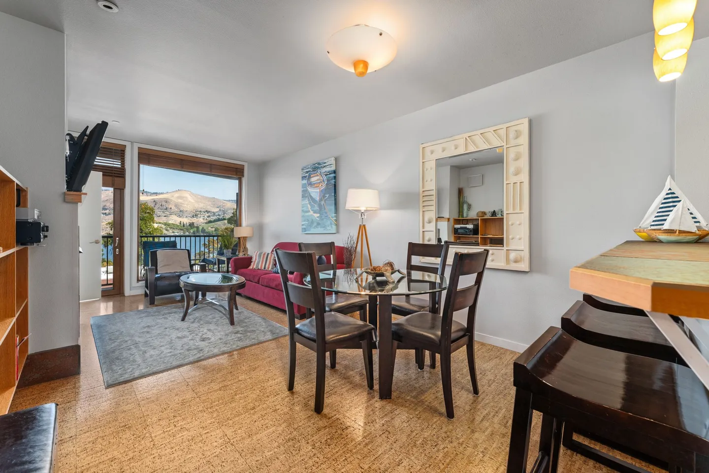 Open concept dining and living area featuring a round glass-top dining table, a red couch, and sliding glass doors with a view of a lake and hills.