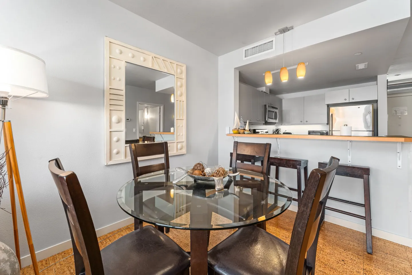 Dining area with a round glass-top table, dark wood chairs, a large decorative framed mirror, and a view into the kitchen pass-through counter.