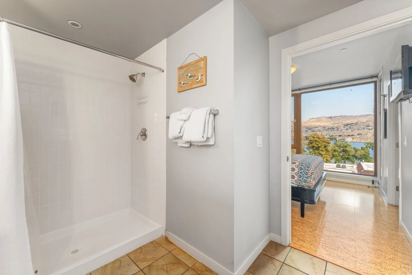 Bathroom with a tiled walk-in shower, folded towels on a rack, and an open doorway leading to a sunlit bedroom with scenic water views.