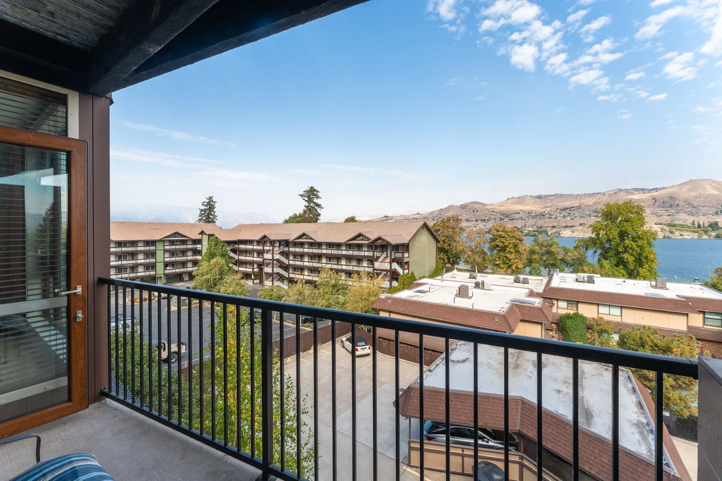 View from an upper-level balcony with a black metal railing overlooking condo buildings, parking, and a lake with hills in the distance.