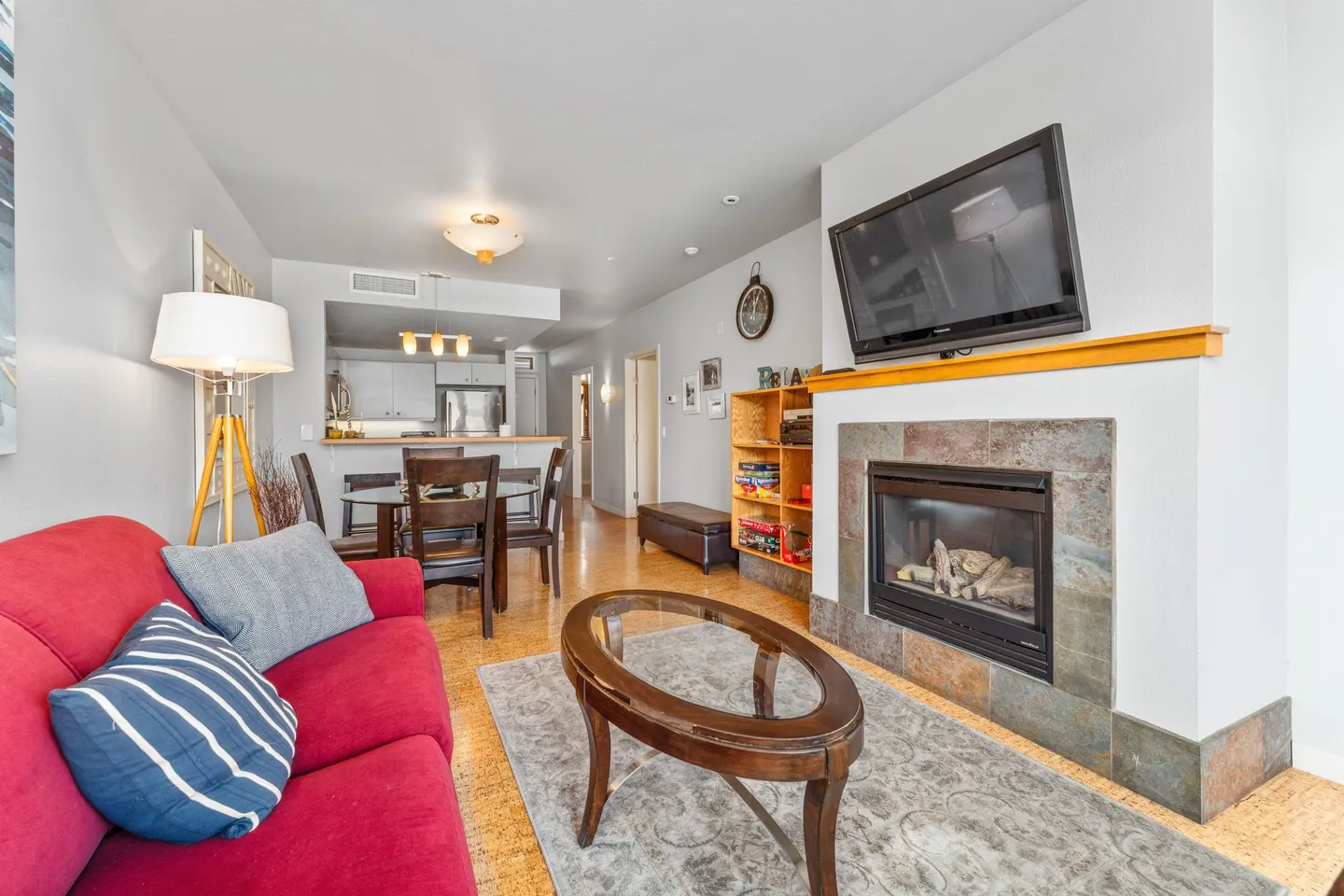 Open-concept living room featuring a red couch, tiled fireplace with mounted TV, oval coffee table, dining set, and kitchen in the background.