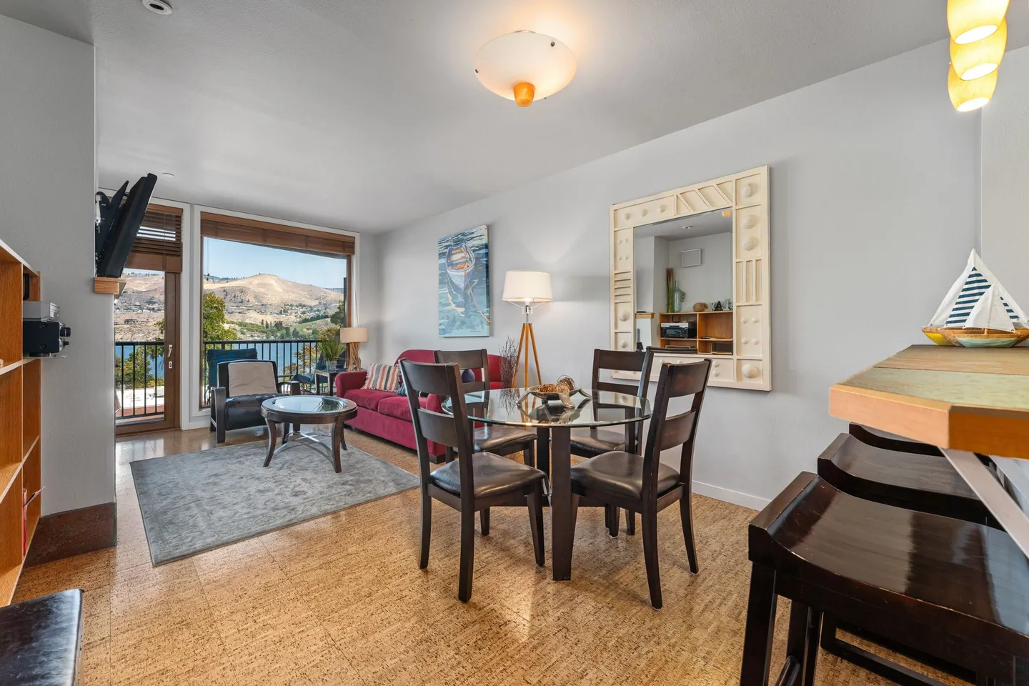 Open living and dining area featuring a round glass dining table with chairs, patterned flooring, a living room with a red sofa, and large windows looking out to a body of water and hills.