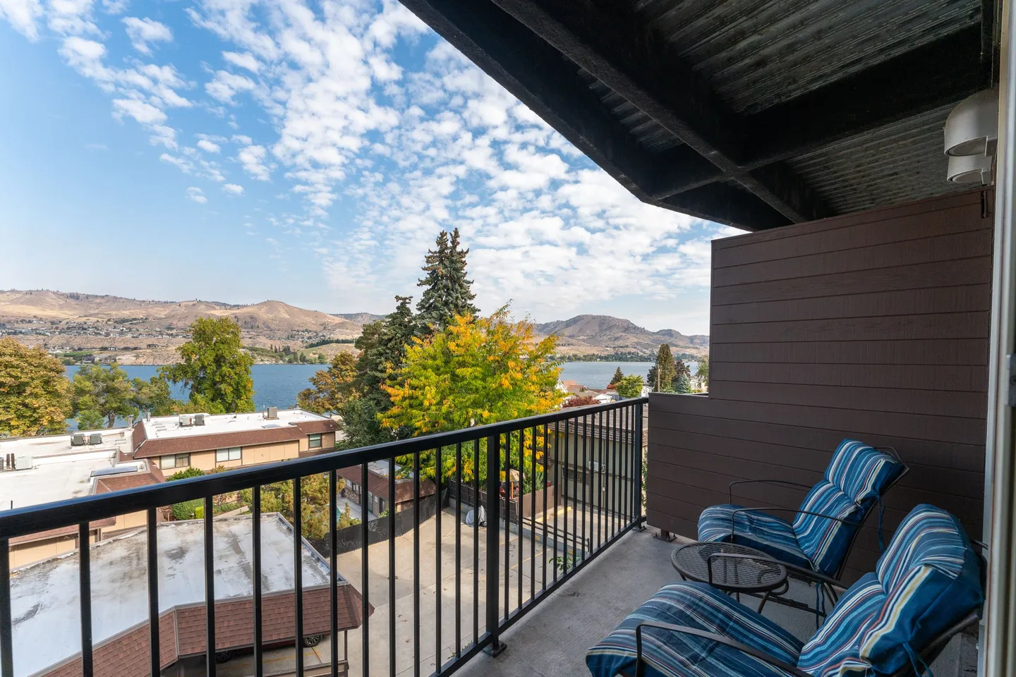 Balcony with two blue striped chairs overlooking trees, a lake, and distant hills under a partly cloudy sky.