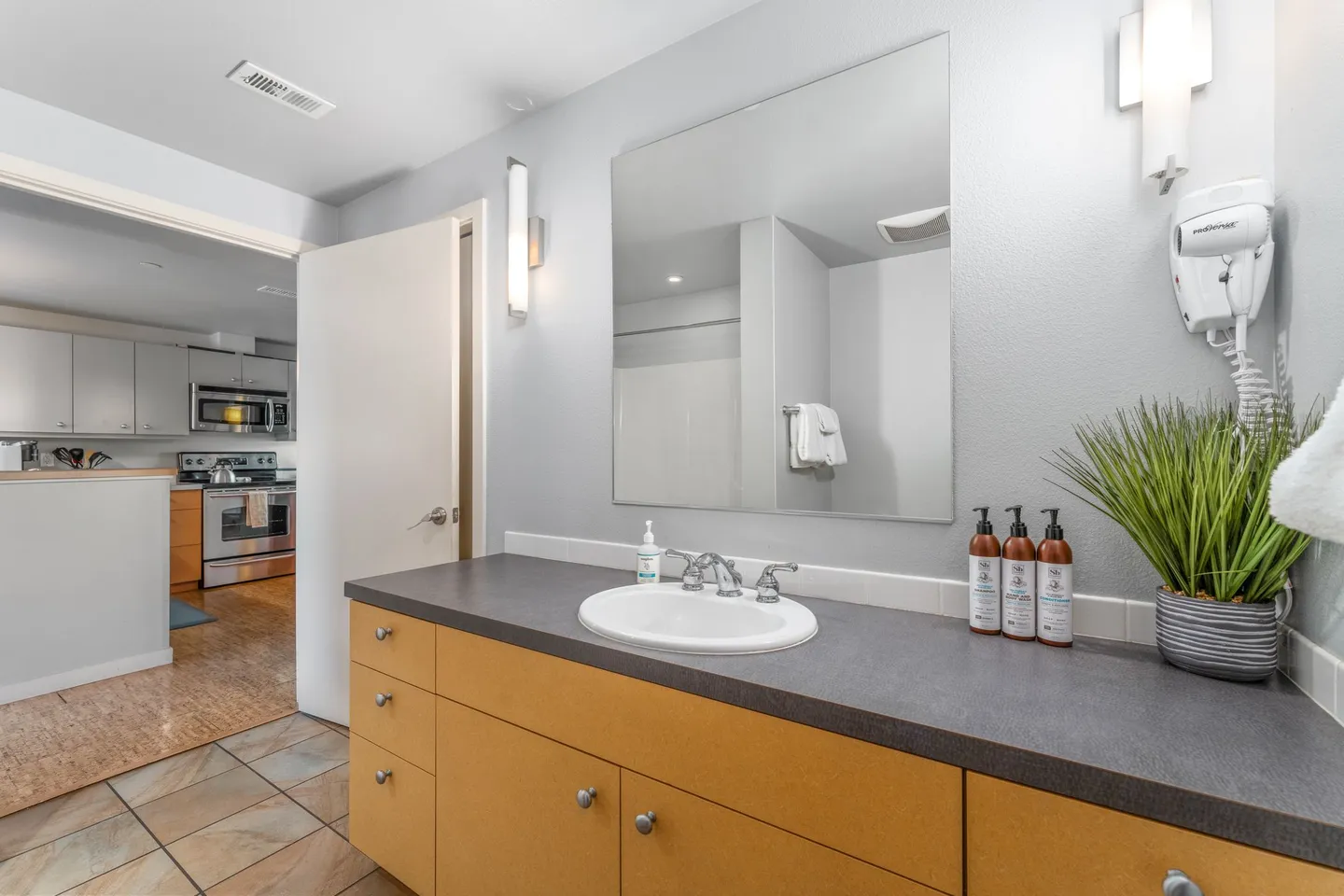Bathroom vanity with a dark countertop, white oval sink, frameless mirror, and light wood cabinetry next to an open doorway showing the kitchen.