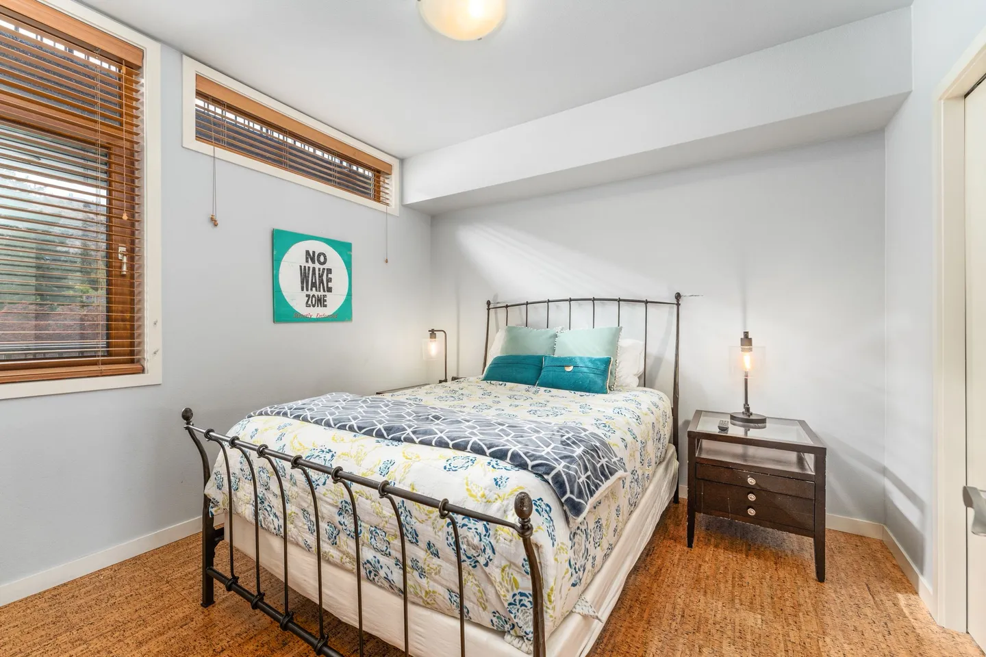 Bedroom featuring a black metal bed frame with patterned bedding, a dark nightstand with a lamp, and high horizontal windows with blinds.