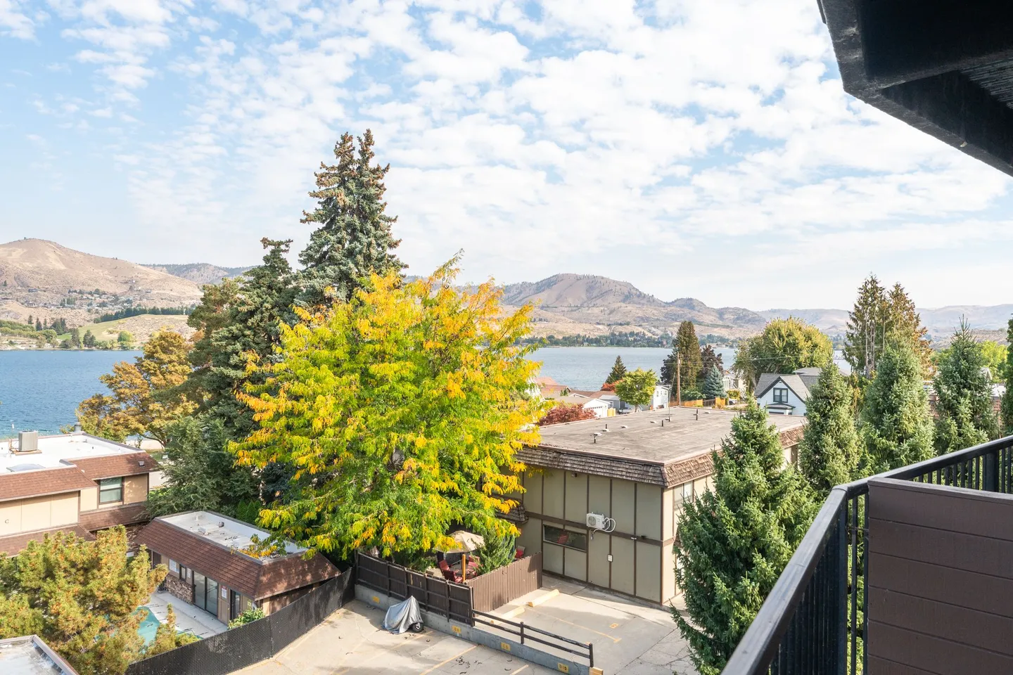 Balcony view overlooking nearby rooftops, trees, a lake, and distant mountains under a blue sky with light clouds.