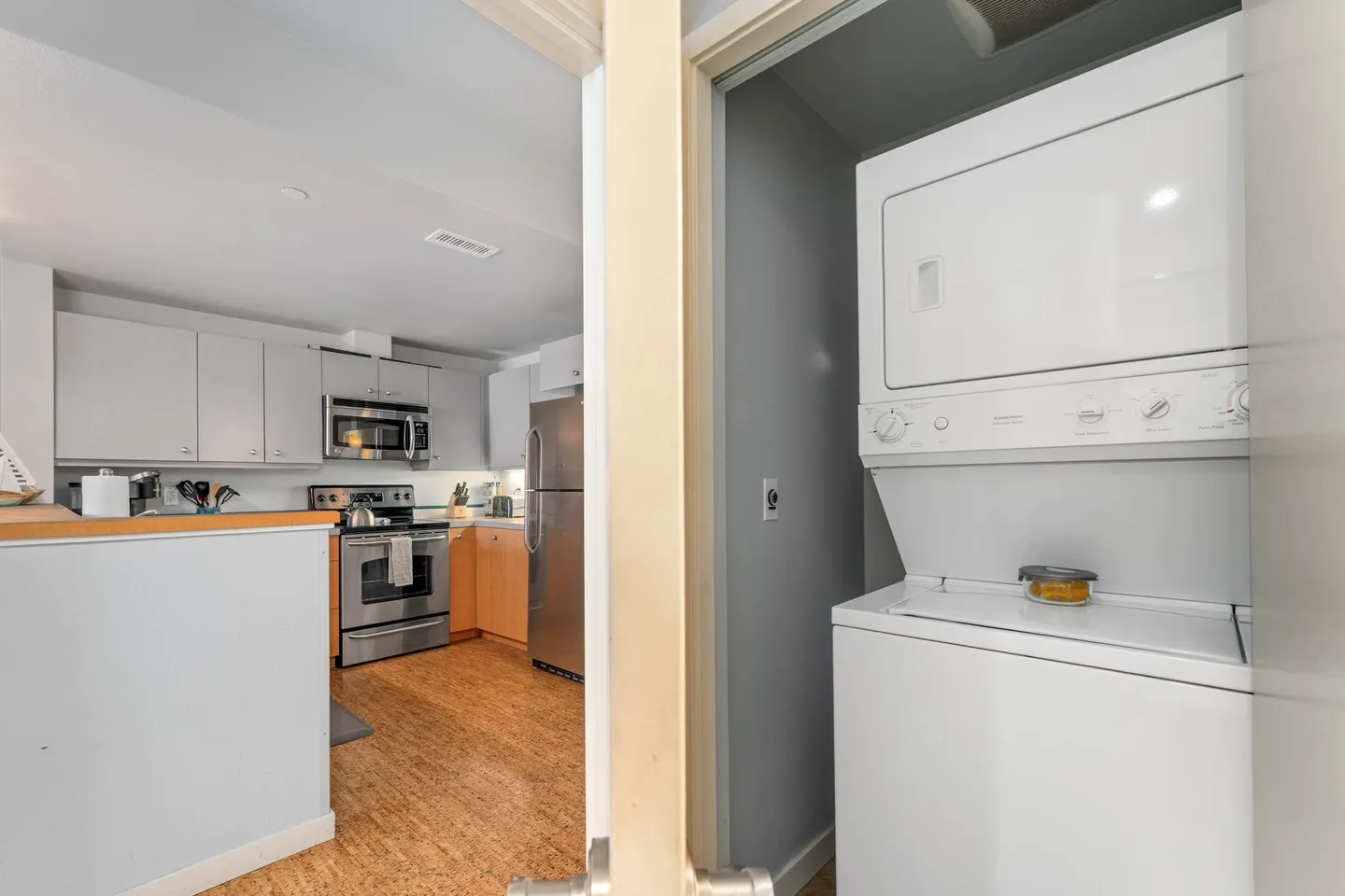 A stacked white washer and dryer unit in a closet, with a view looking into an adjoining kitchen featuring stainless steel appliances.