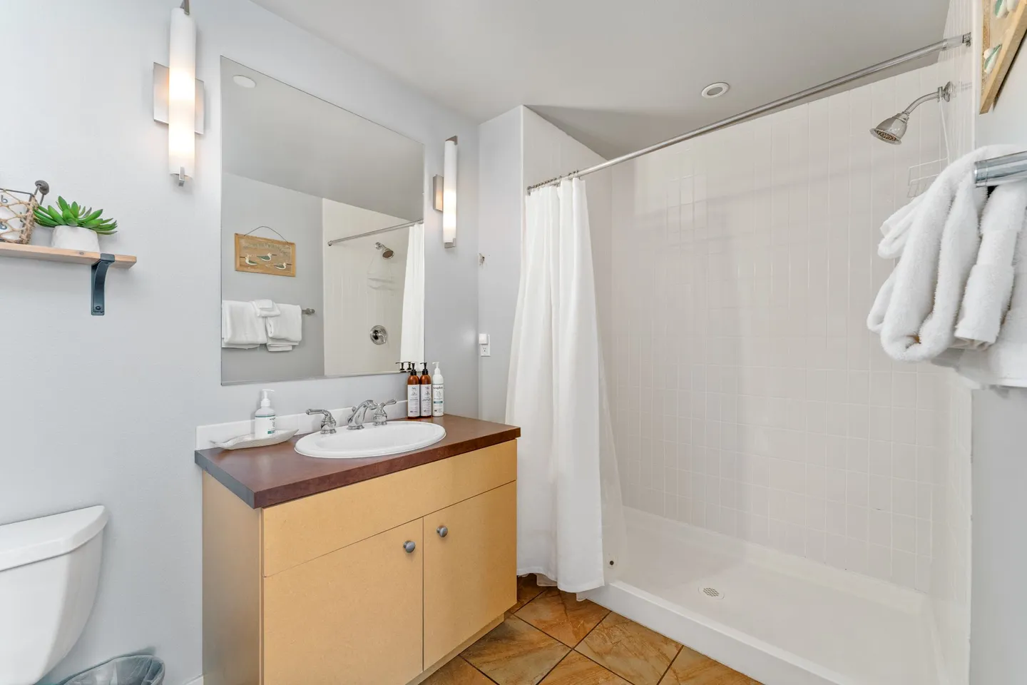 Bathroom featuring a vanity with wood-tone cabinet, a mirror flanked by wall sconces, and a walk-in shower with a white curtain and tiled walls.