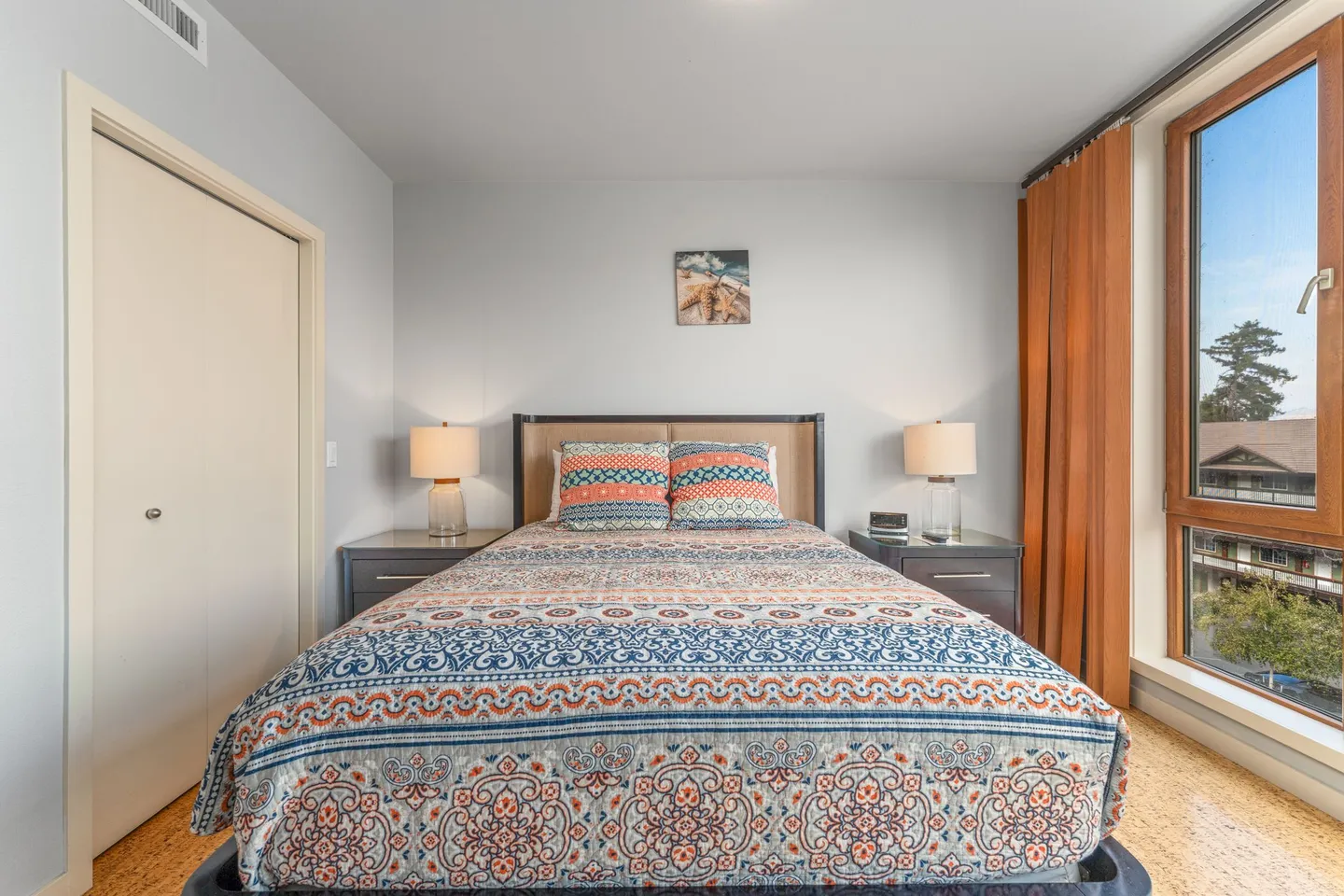 Bedroom with a patterned bedspread, two nightstands with lamps, and a window with orange curtains.