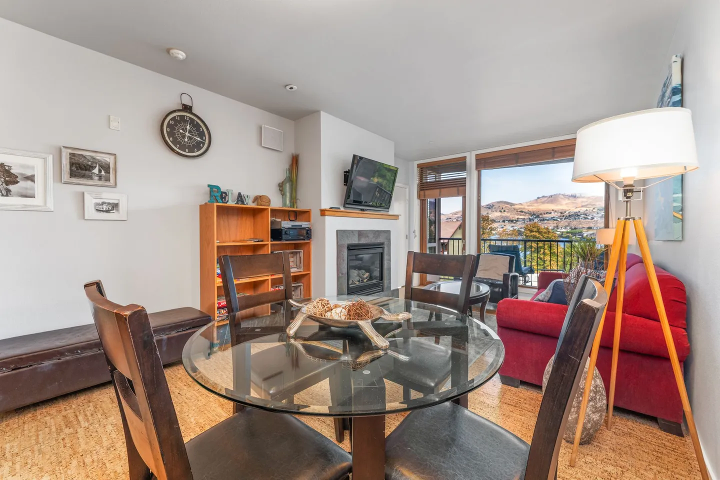 Dining area with a round glass table and chairs, adjacent to a living area with a fireplace, TV, and balcony doors showing mountain views.