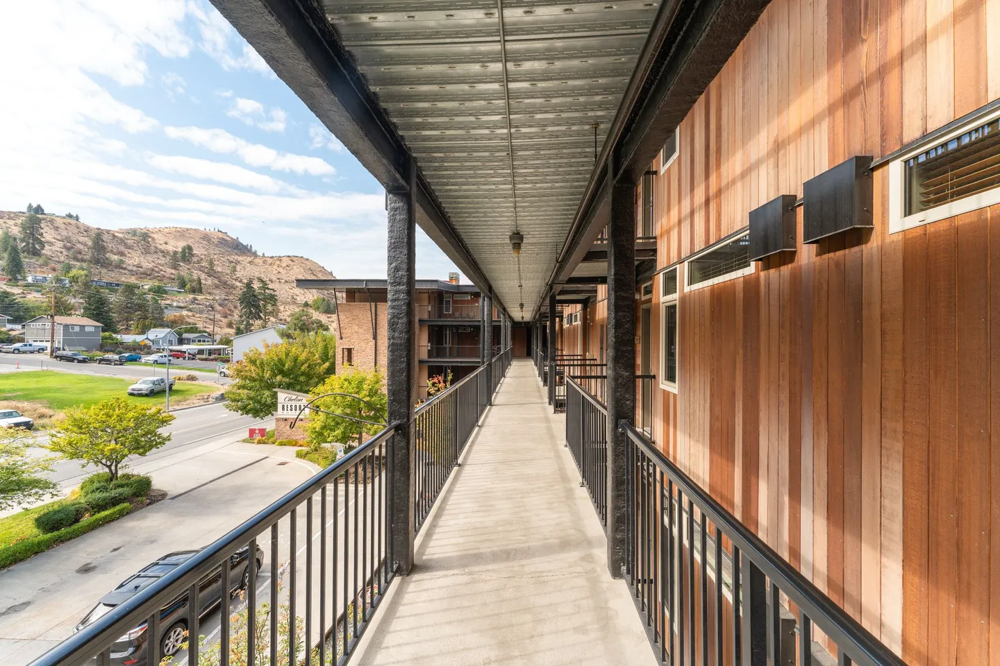 Exterior covered walkway along an apartment or condominium building with black railings and wood-paneled walls.