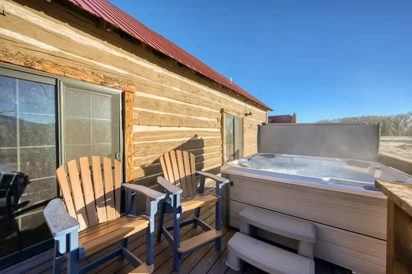 Outdoor wooden deck outside a log cabin featuring two wooden armchairs and a bubbling hot tub under a clear blue sky.