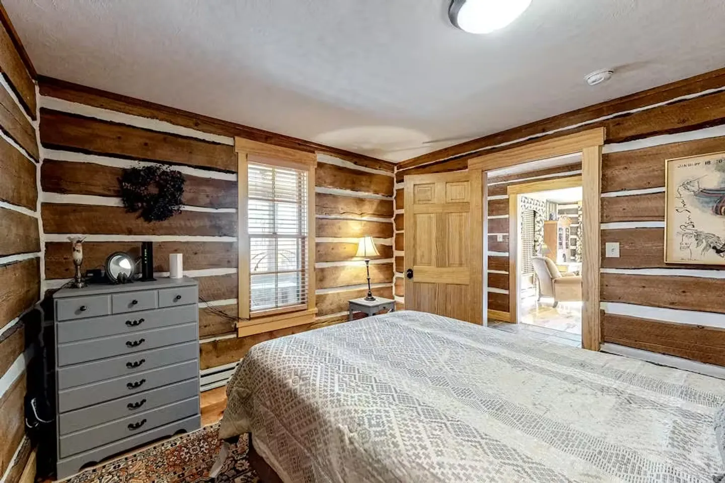 Bedroom with exposed log walls, a bed with a patterned coverlet, a grey dresser, and a window.