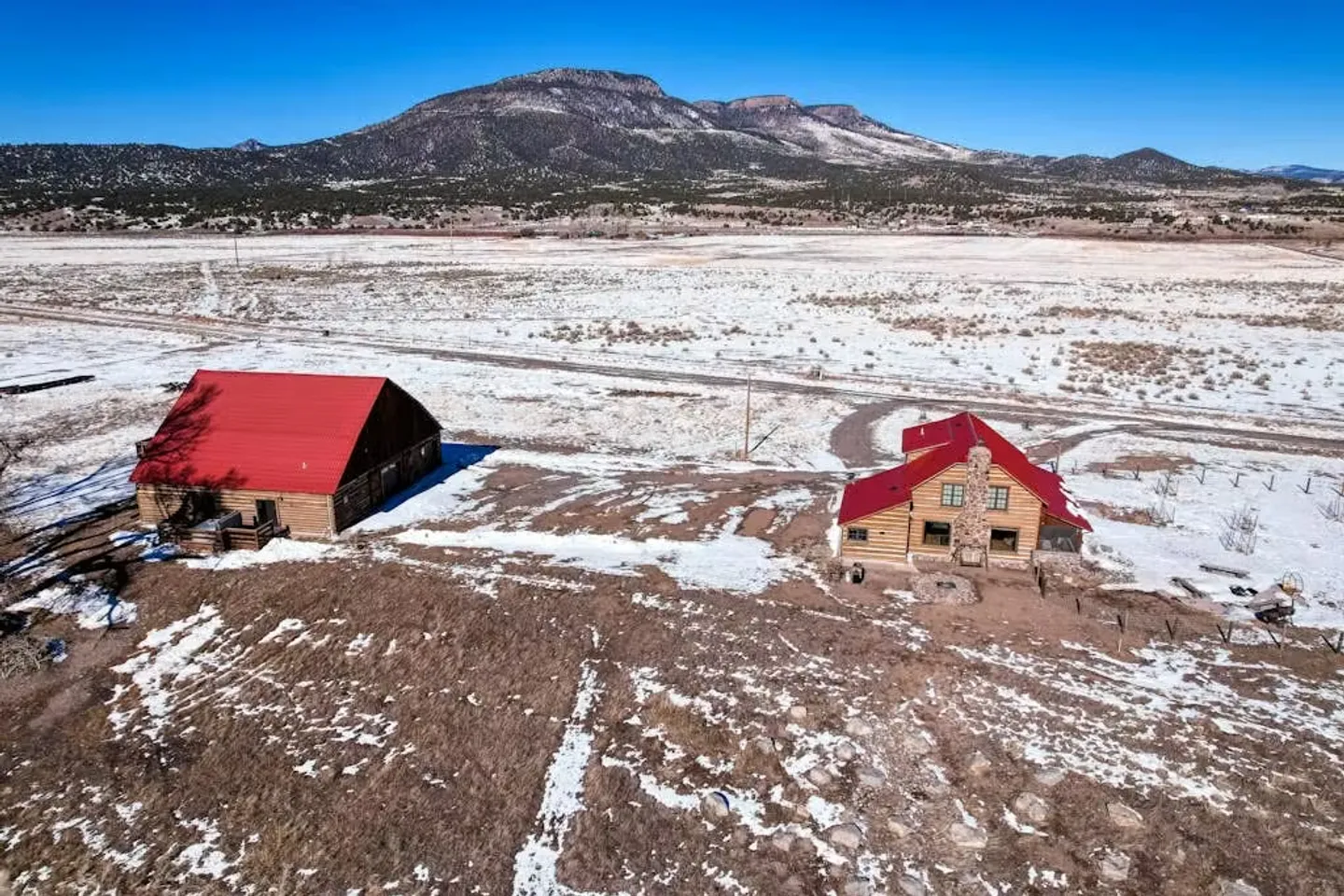 Aerial view of a log cabin and a red-roofed barn in a snowy rural landscape with mountains in the background under a blue sky.