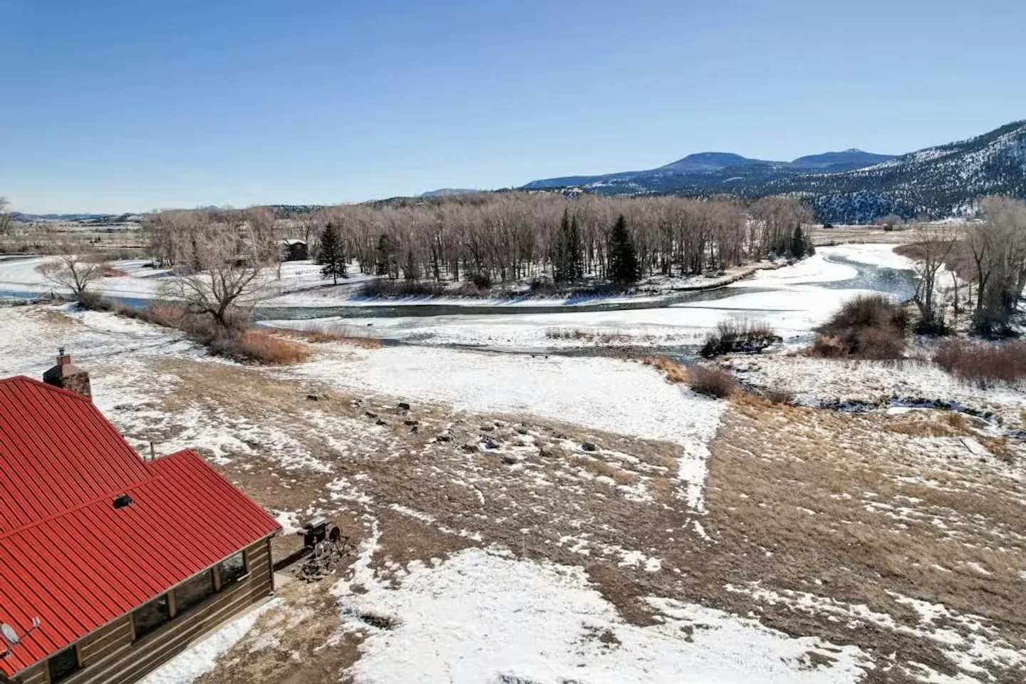 Aerial view of a cabin with a red metal roof overlooking a snow-dusted landscape and a winding river.