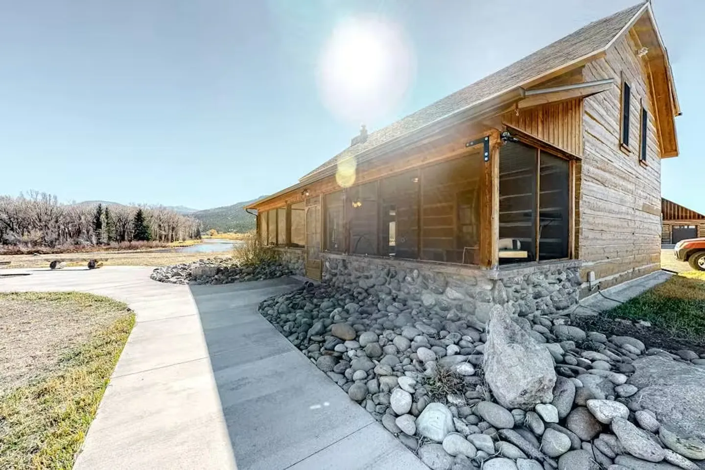 Exterior view of a wood and stone cabin with a screened porch, paved walkway, river rock landscaping, and a distant body of water.