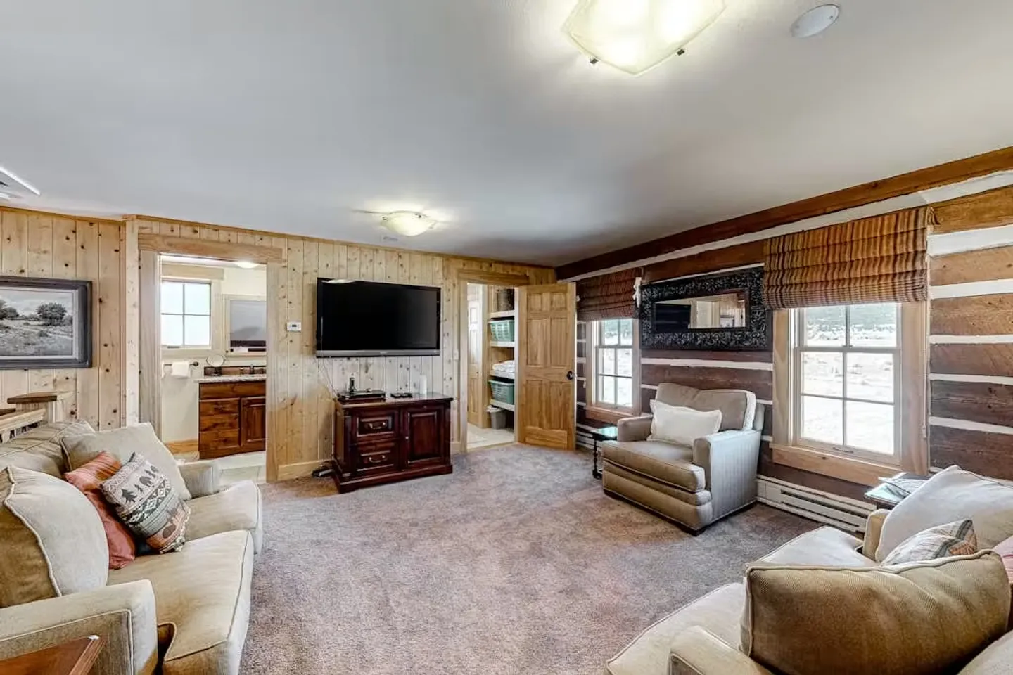 Living room with carpeted floor, log and wood-paneled walls, comfortable armchairs, a couch, and a wall-mounted TV.