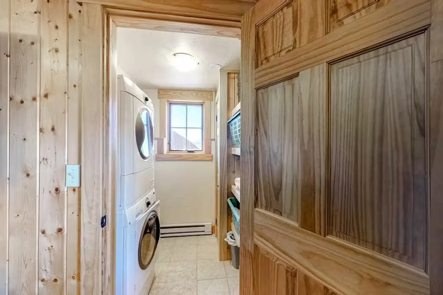 A view through a wooden doorway into a compact laundry room with a stacked white washer and dryer, tiled flooring, and a window.