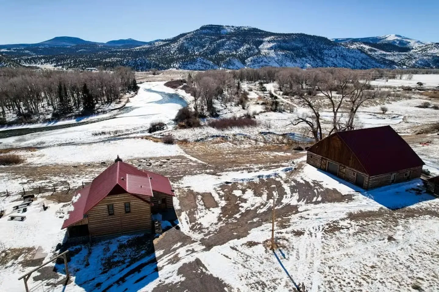 Aerial view of a log cabin and a barn with red roofs situated in a snow-dusted valley near a river and mountains.
