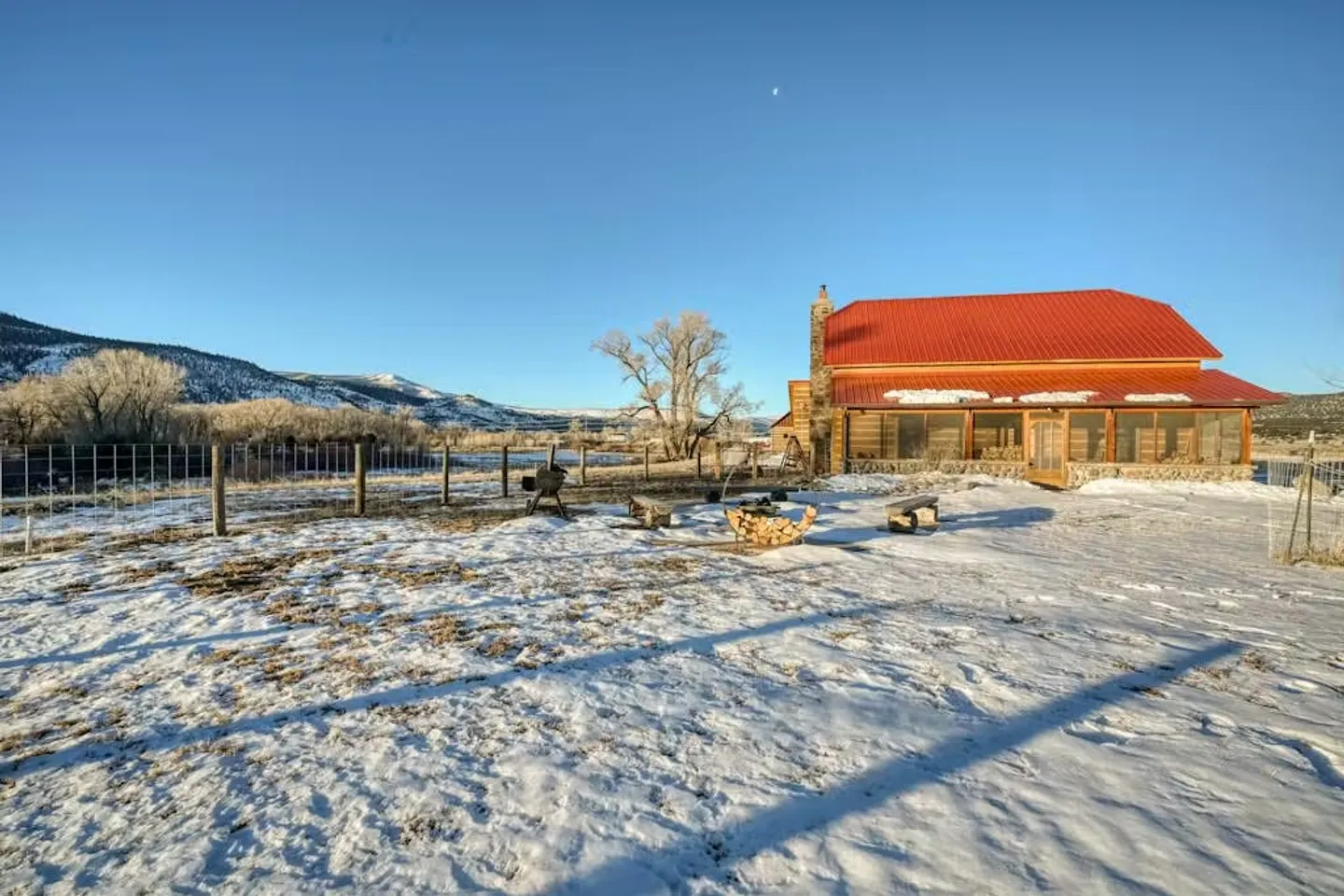 Exterior view of a cabin with a red metal roof surrounded by a snow-covered yard, fence, and mountain landscape.