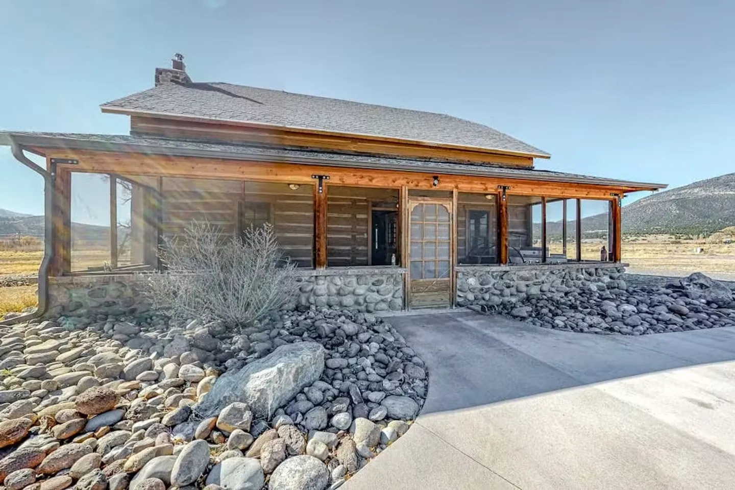 Exterior view of a rustic log cabin with a river-rock foundation, screened porch, and surrounding rock landscaping under a clear sky.