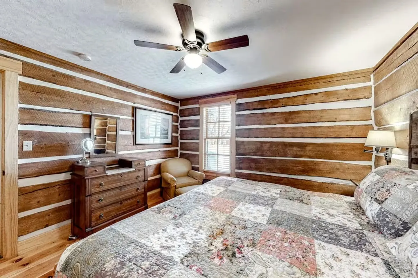 Bedroom with log walls, chinking, a patchwork quilt on the bed, a wooden dresser, armchair, and ceiling fan.