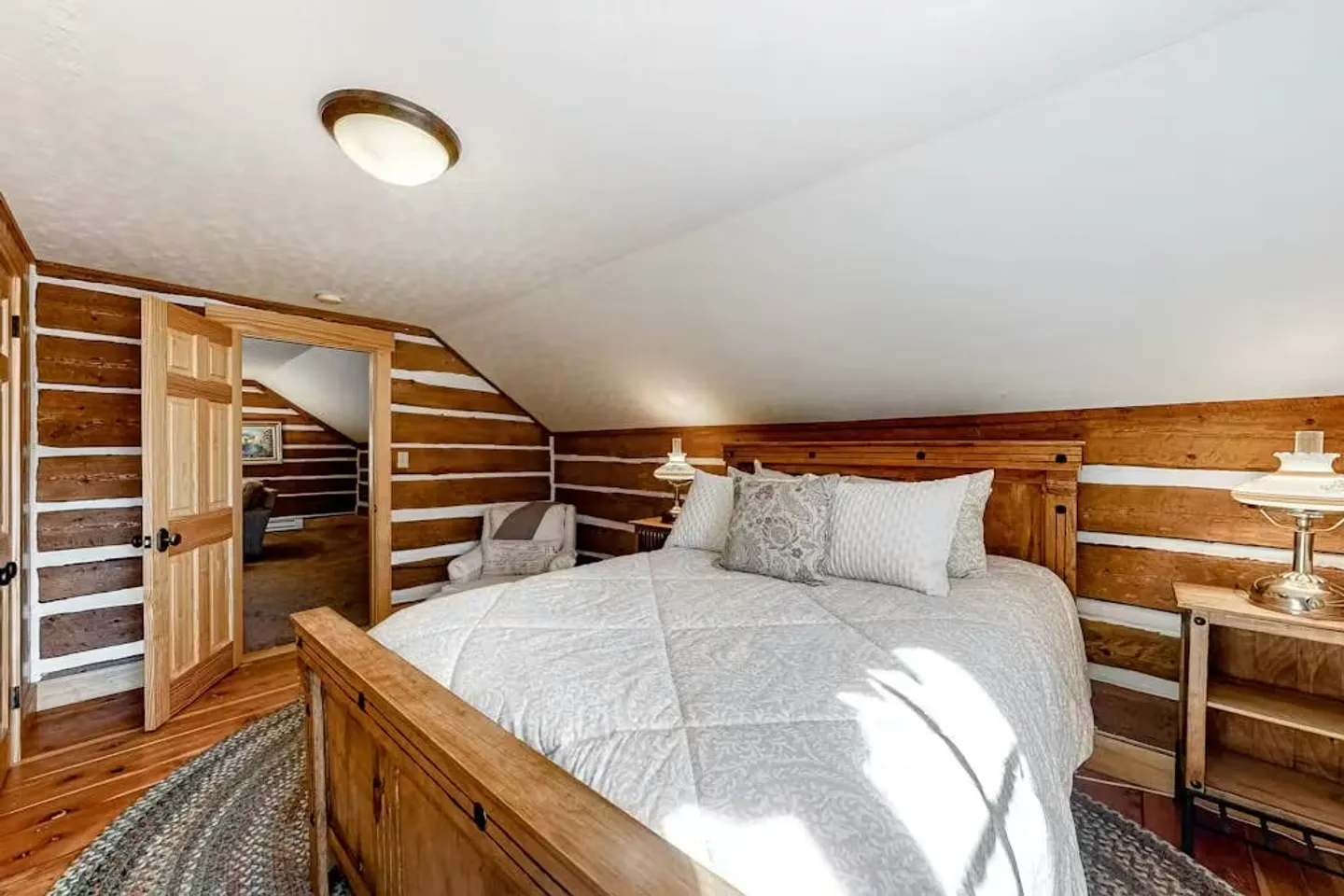 Bedroom with log walls, a sloped ceiling, wooden bed frame, bedside tables with lamps, and an open doorway leading to another room.