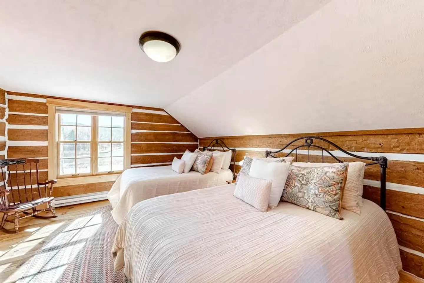 Upstairs log-walled bedroom with two beds, an angled ceiling, a window, and a wooden rocking chair.