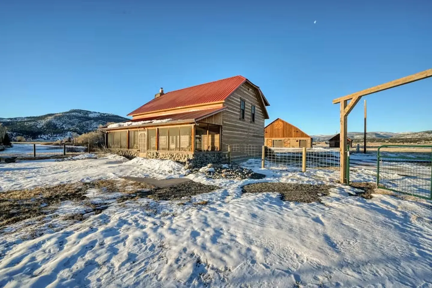 Exterior of a two-story log cabin with a red metal roof and enclosed porch set in a snowy landscape with distant mountains under a blue sky.